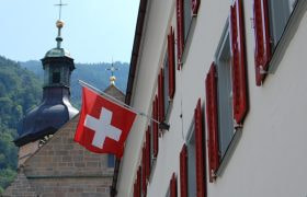 Bandiera svizzera su edificio con chiesa alpina sullo sfondo.