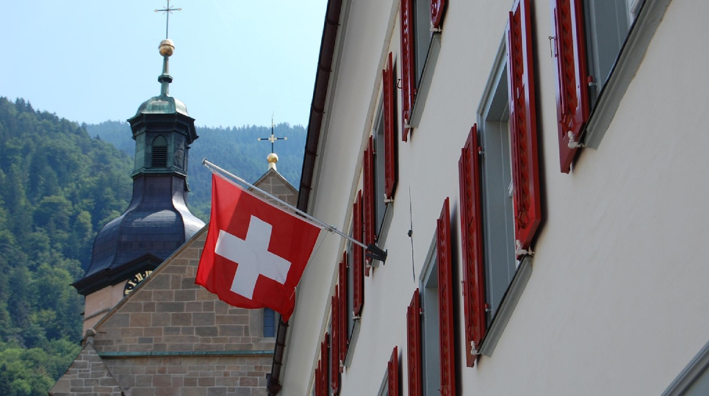 Bandiera svizzera su edificio con chiesa alpina sullo sfondo.