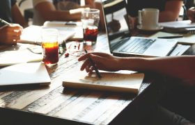 Persone al lavoro con laptop, taccuini e bevande.