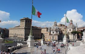Piazza romana con bandiere italiane, cupole storiche ed escursionisti in visita.