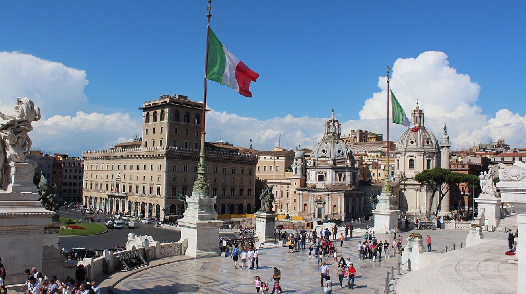 Piazza romana con bandiere italiane, cupole storiche ed escursionisti in visita.