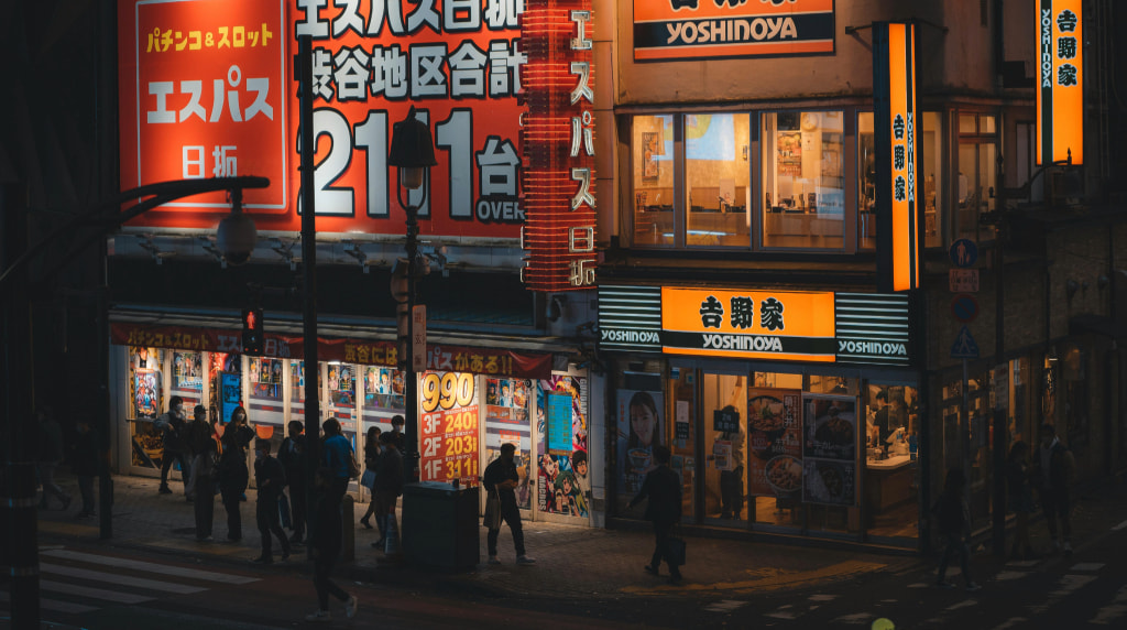 Strada giapponese con insegne luminose, locali affollati e passanti notturni.
