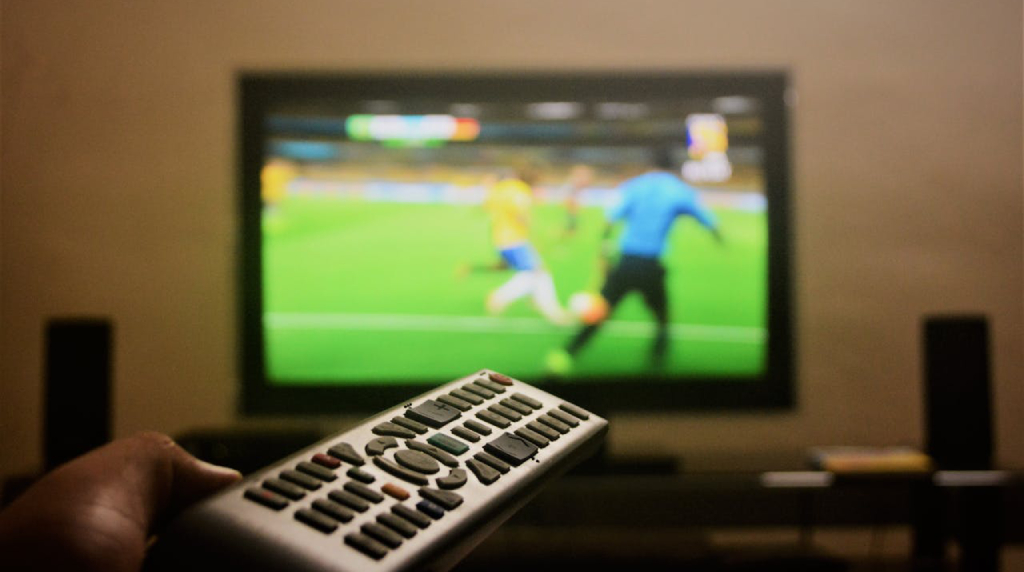 Televisie met voetbalwedstrijd en een hand die afstandsbediening vasthoudt