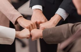 Patru mâini care își ciocnesc pumnii într-un gest de colaborare, în fundal roți dințate colorate.