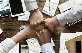 Patru persoane își unesc pumnii deasupra unei mese de birou.