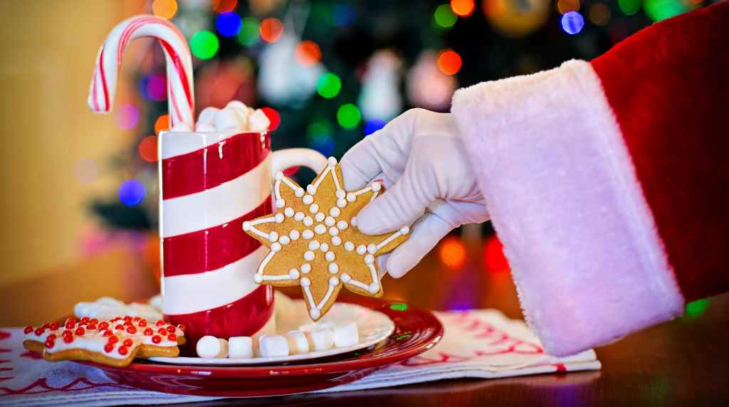 Mâna lui Moș Crăciun ridicând o turtă dulce lângă o cană festivă cu bețe de zahăr
