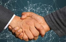 Two people in suits shaking hands in front of a background with a digital graphic design on it.
