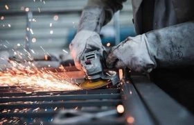 A worker using a blade saw on a piece of metal in a factory, with sparks flying out from the blade grinding against the metal.