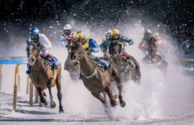 Several jockeys racing their horses at full gallop in the snow on an outdoor horse racing track.