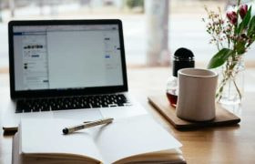 An open notebook lying flat on a table in front of a laptop next to a coffee mug and flower vase.