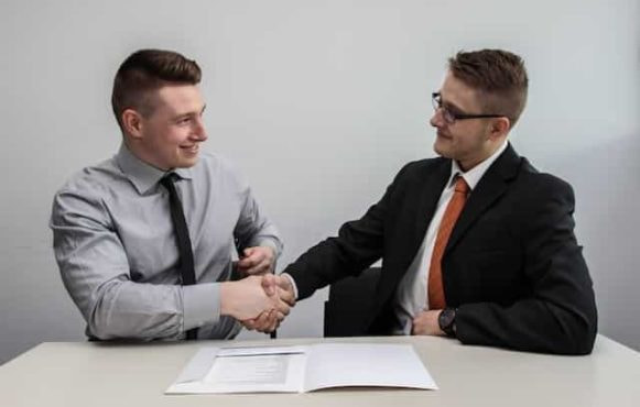 Two men in suits sitting down and facing one another while shaking each others’ hands.