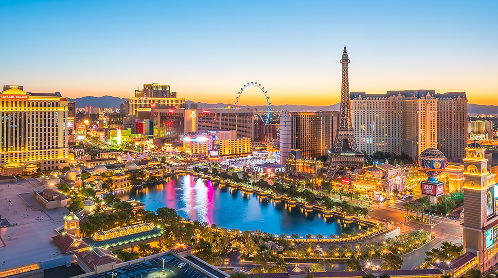 View of the Las Vegas Strip at sunset, featuring illuminated hotels, replicas likethe Eiffel Tower, and a large fountain-lined lagoon.