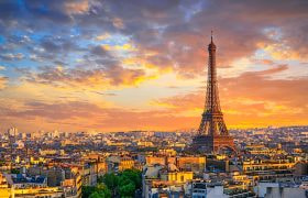Eiffel Tower overlooking Paris at sunset, with warm clouds and city buildings below.