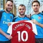 Three Arsenal players holding up a club shirt with the name CashBet Coin printed on it.