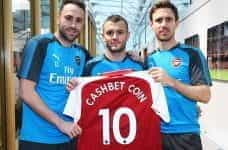 Three Arsenal players holding up a club shirt with the name CashBet Coin printed on it.