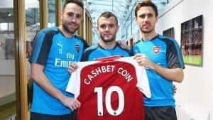 Three Arsenal players holding up a club shirt with the name CashBet Coin printed on it.