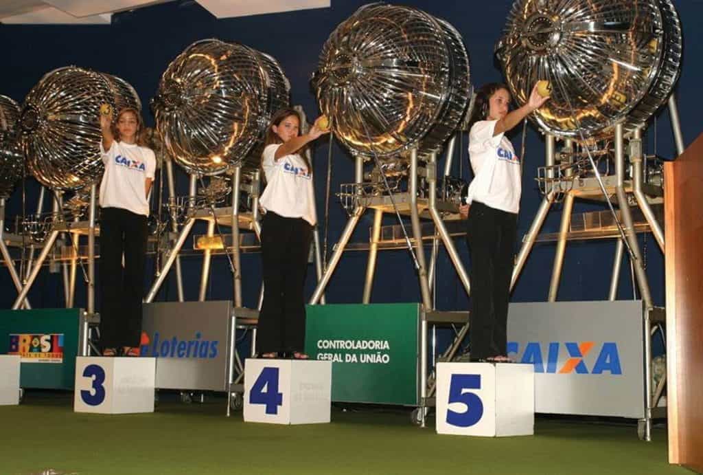 Three women stand on cubes to pick numbers from large metallic wheels to randomly select several winning lottery numbers.