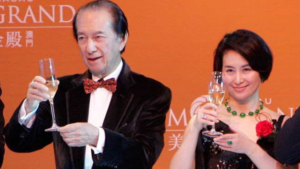 Pansy and Stanley Ho, dressed in black tie attire, raise champagne glasses at an event in front of an orange backdrop.