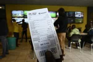 A hand holds up a betting slip in front of rows of illuminated televisions.