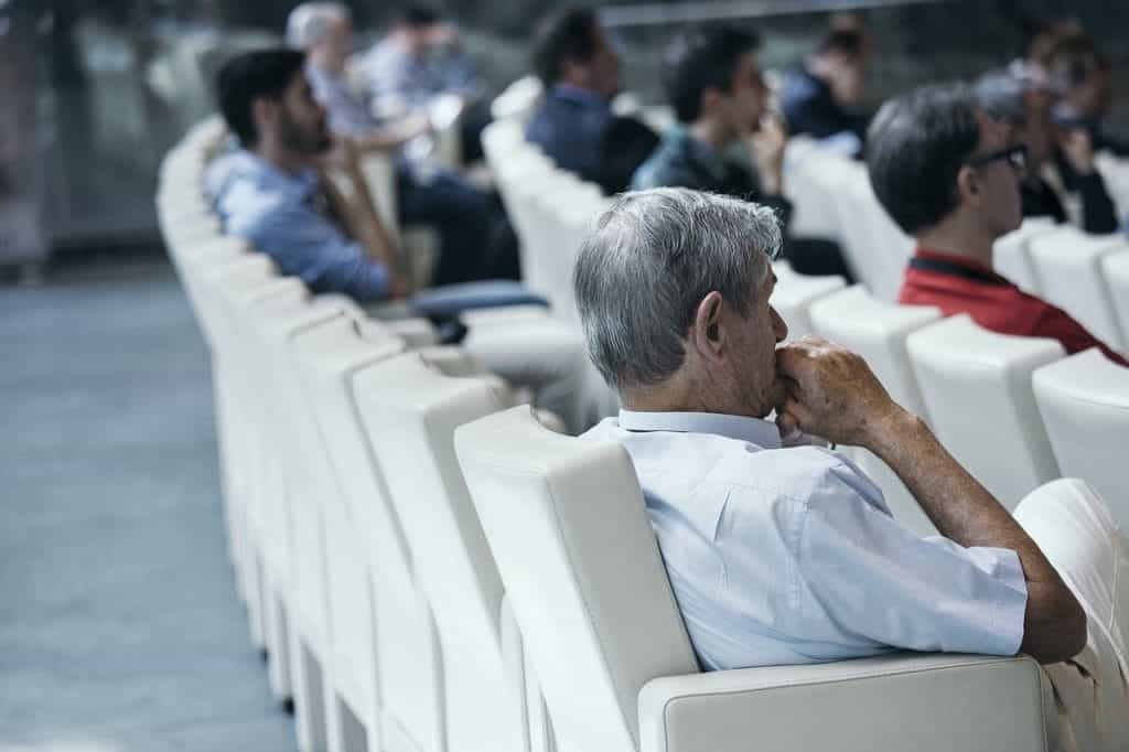 An audience sits in white theatre seats, listening closely.