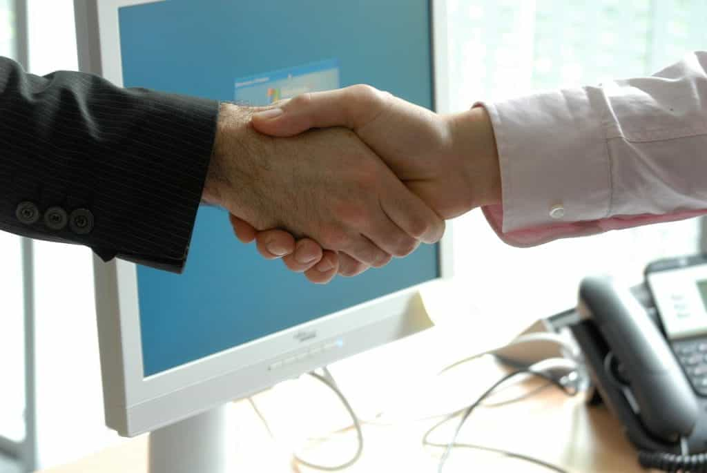 Two white hands shake in front of a computer screen.