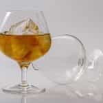 A glass of brandy and ice stands while another empty glass is tipped over behind it.