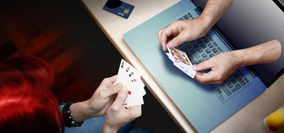 A person holding a hand of cards in front of a laptop, with a separate pair of hands coming out of the computer screen, holding another hand of cards.