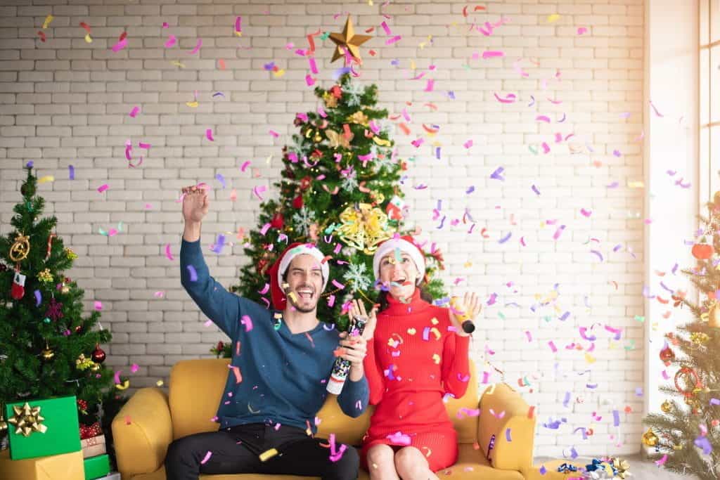 A man and a woman sit on a sofa celebrating Christmas, with decorated trees in the background and colorful confetti falling down.