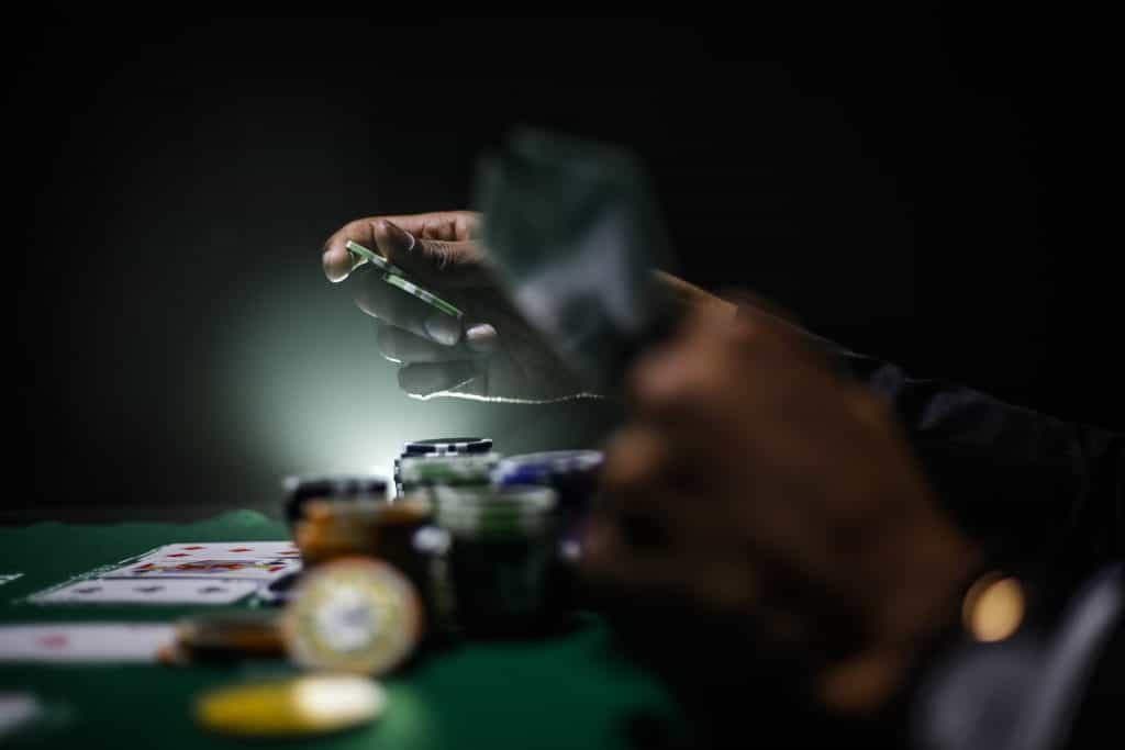 A dark, sideways look down a poker table at hands playing poker: one set holding up cards, the other setting down two green chips to place a bet.