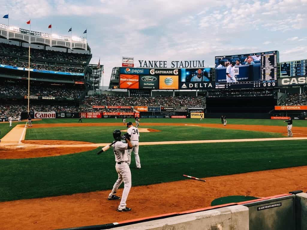 A full crowd at Yankee Stadium watches baseball, with a batter warming up.