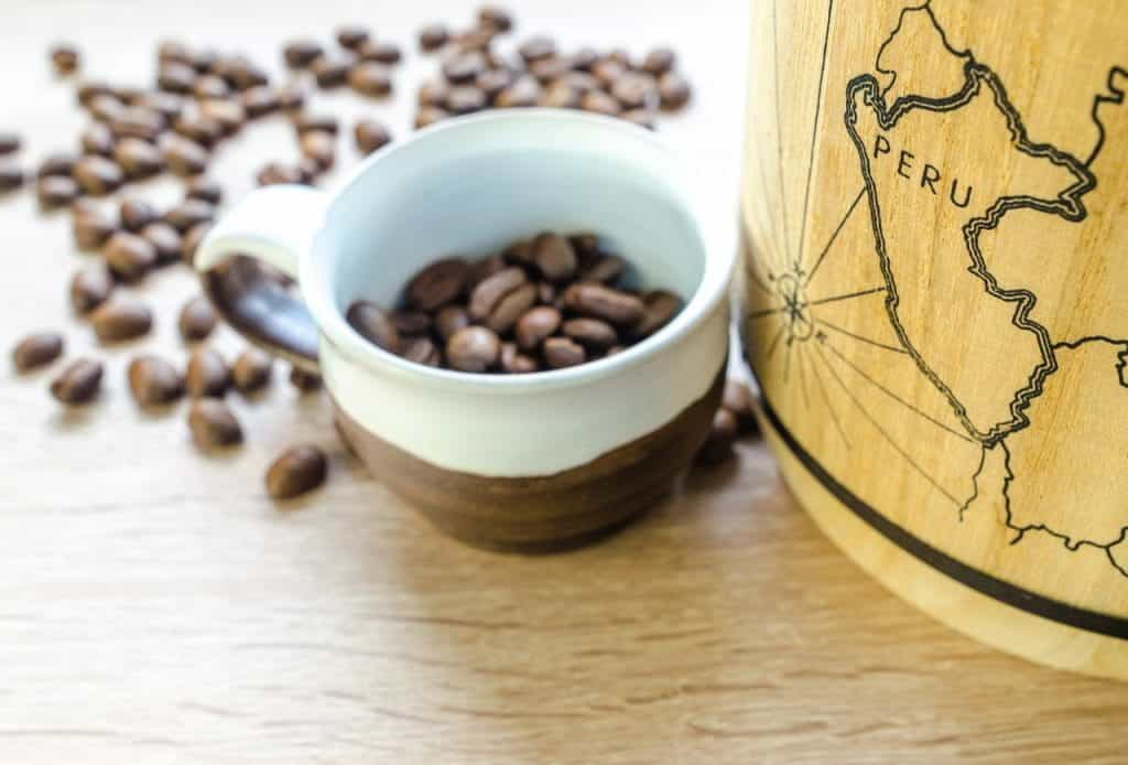 A small mug filled with coffee beans sits next to a map printed on wood that marks the location of Peru.