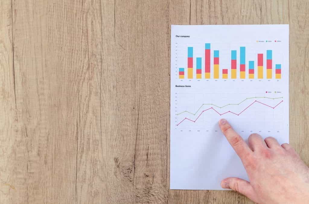 A look down at a wooden desk, where a white hand points to a line graph demonstrating an upward trend.