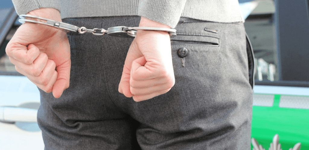 A pair of white hands handcuffed behind someone’s back in front of a grey police car.