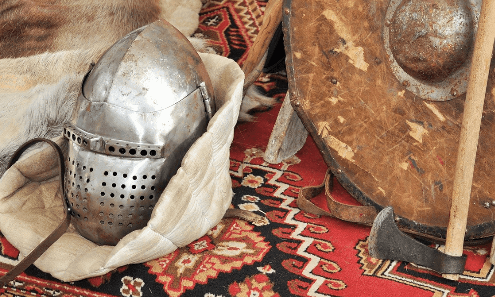 A colorful look down at an array of medieval armory on a red rug: a shield, helmet, axe, and fur.