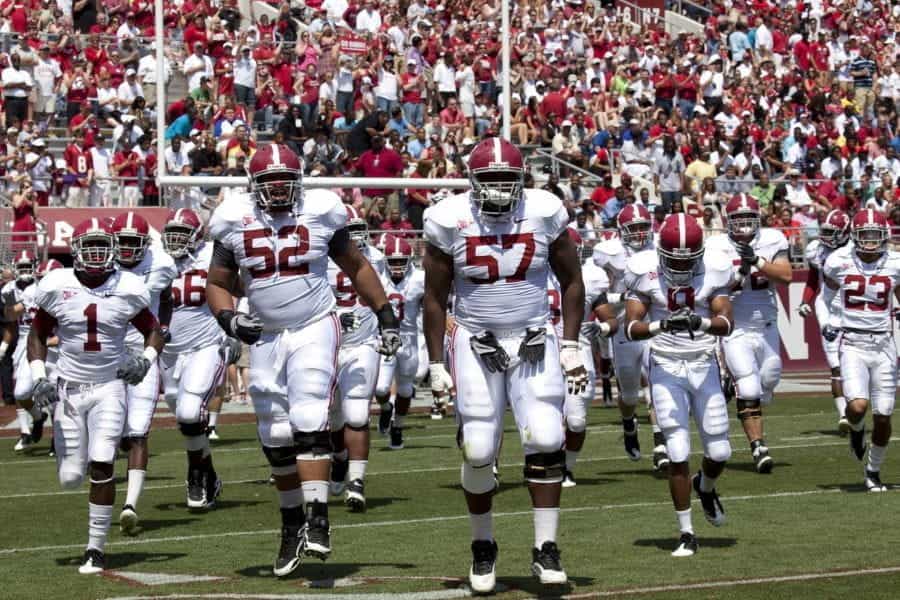 Football players in white and red uniforms stand on the field while a stadium full of fans watch behind them.