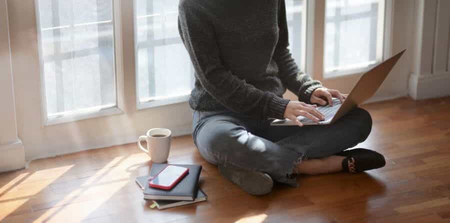 A woman sits in a window with a laptop, coffee and notebooks.