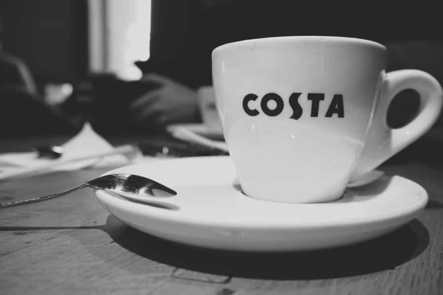 A black-and-white photograph of a white coffee cup reading COSTA.