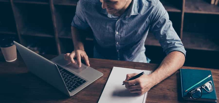 A man taking notes, while looking at a laptop.