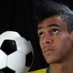 A soccer player wearing a Brazil team jersey holds a soccer ball on his shoulder and looks up.