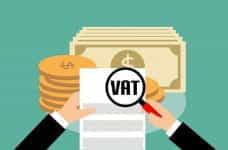 A pair of hands holding a magnifying glass to a piece of paper reading VAT, in front of a pile of money.