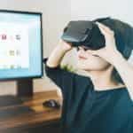 A woman sitting at a computer looks through a set of VR goggles.