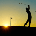 A golf player swings the club to hit a ball against the sunset.