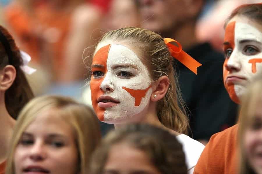 A young sports fan with her face orange and white painted stands in the stands of a sports game.