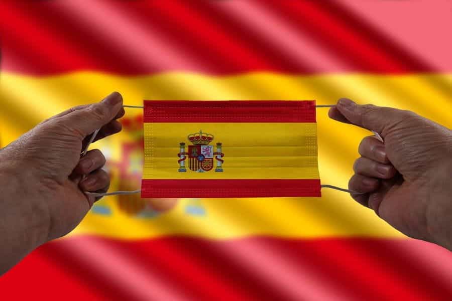 A pair of hands hold a mask with the Spanish flag on it in front of a Spanish flag.