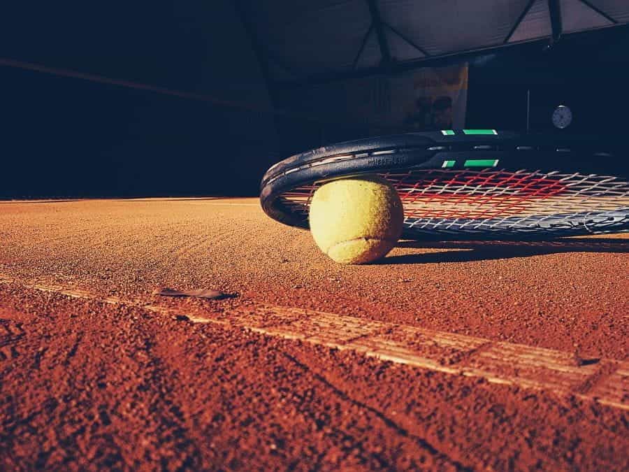 A tennis ball under a racket on a clay court.