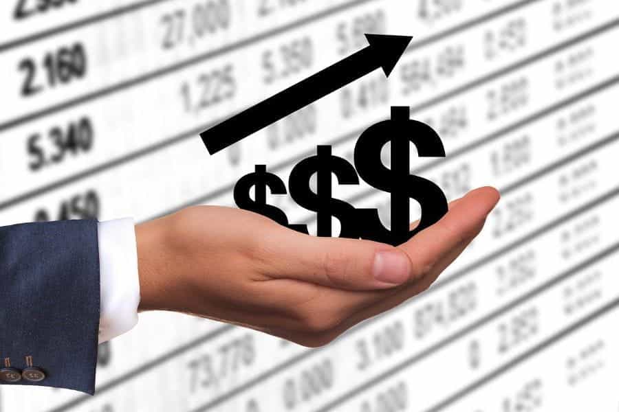 A white hand holds a graphic of three dollar signs in front of a black-and-white image of stock prices.
