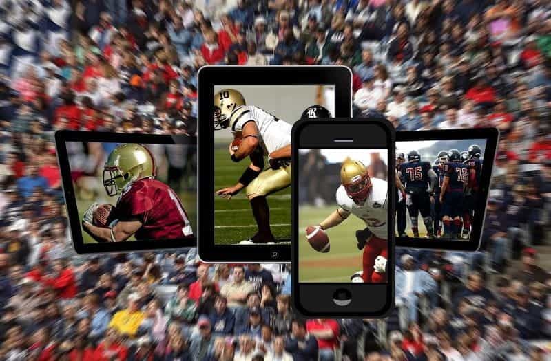 Three digital tablets and a smart phone play football games over a blurred background of sports fans in a stadium.