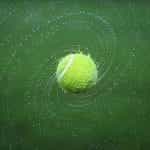 A wet tennis ball in play, flying through the air as its velocity shakes off the water in which it is drenched.