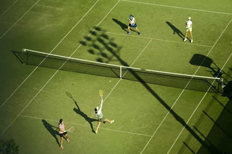 Pertandingan tenis duet wanita di lapangan rumput, dengan matahari yang membuat bayangan dramatis melintasi lapangan.