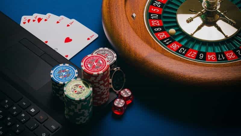 A roulette wheel, cards, poker chips and dice next to a laptop.